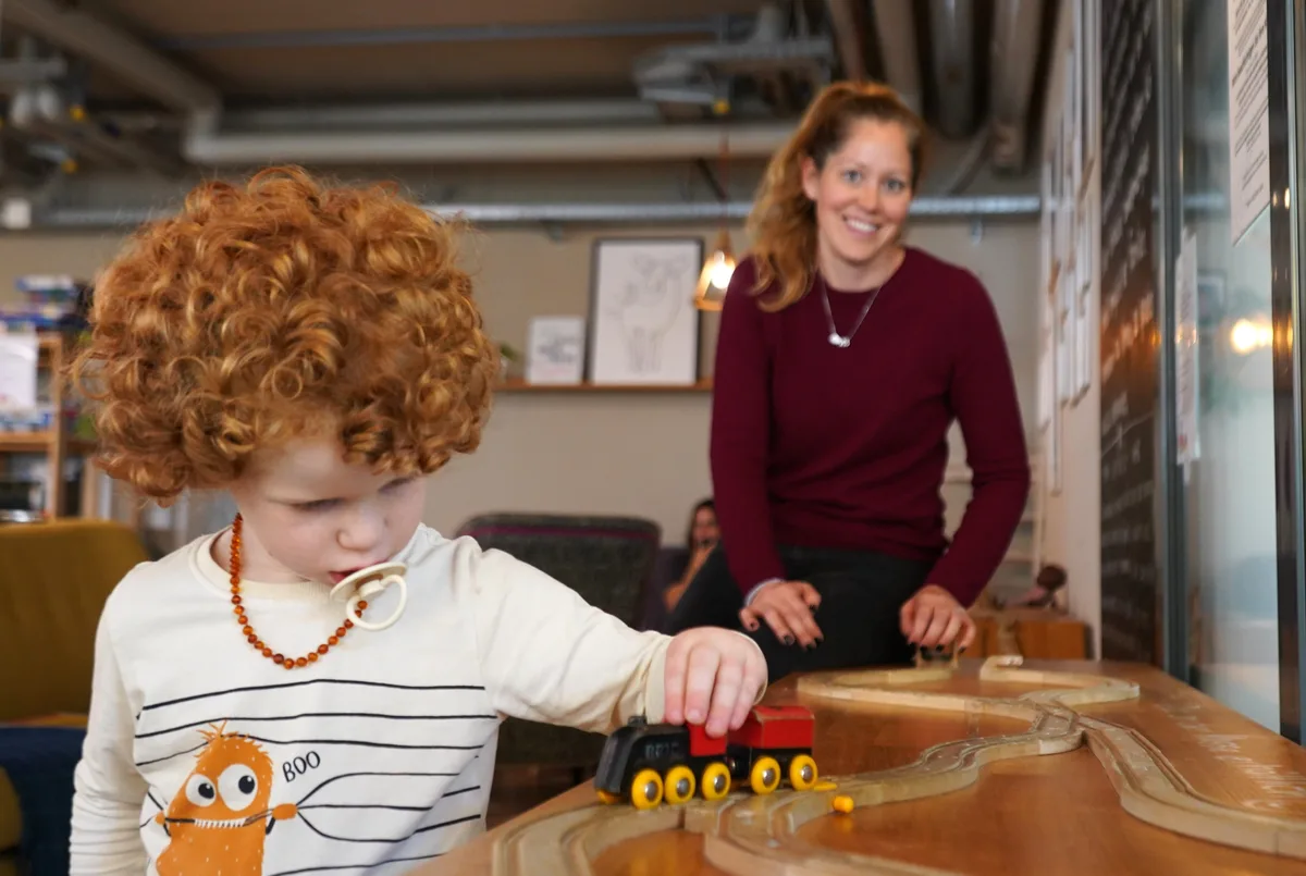 Der 3-jährige Shane fährt eine Lok in der Spielecke des Cafés, im Hintergrund seine Mutter.